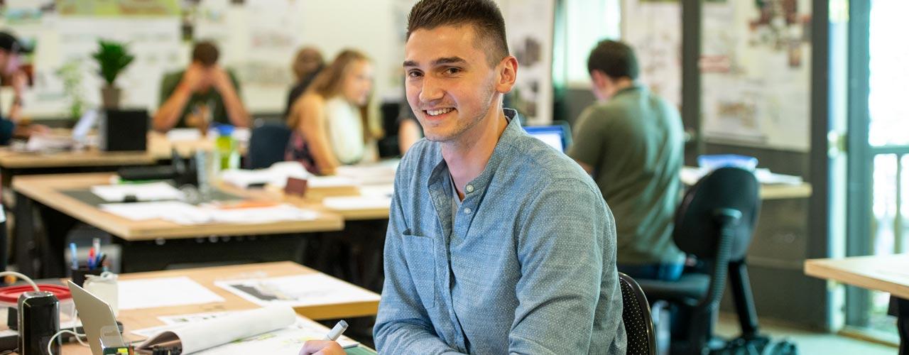 White male college student sitting at desk, looking at camera and smiling